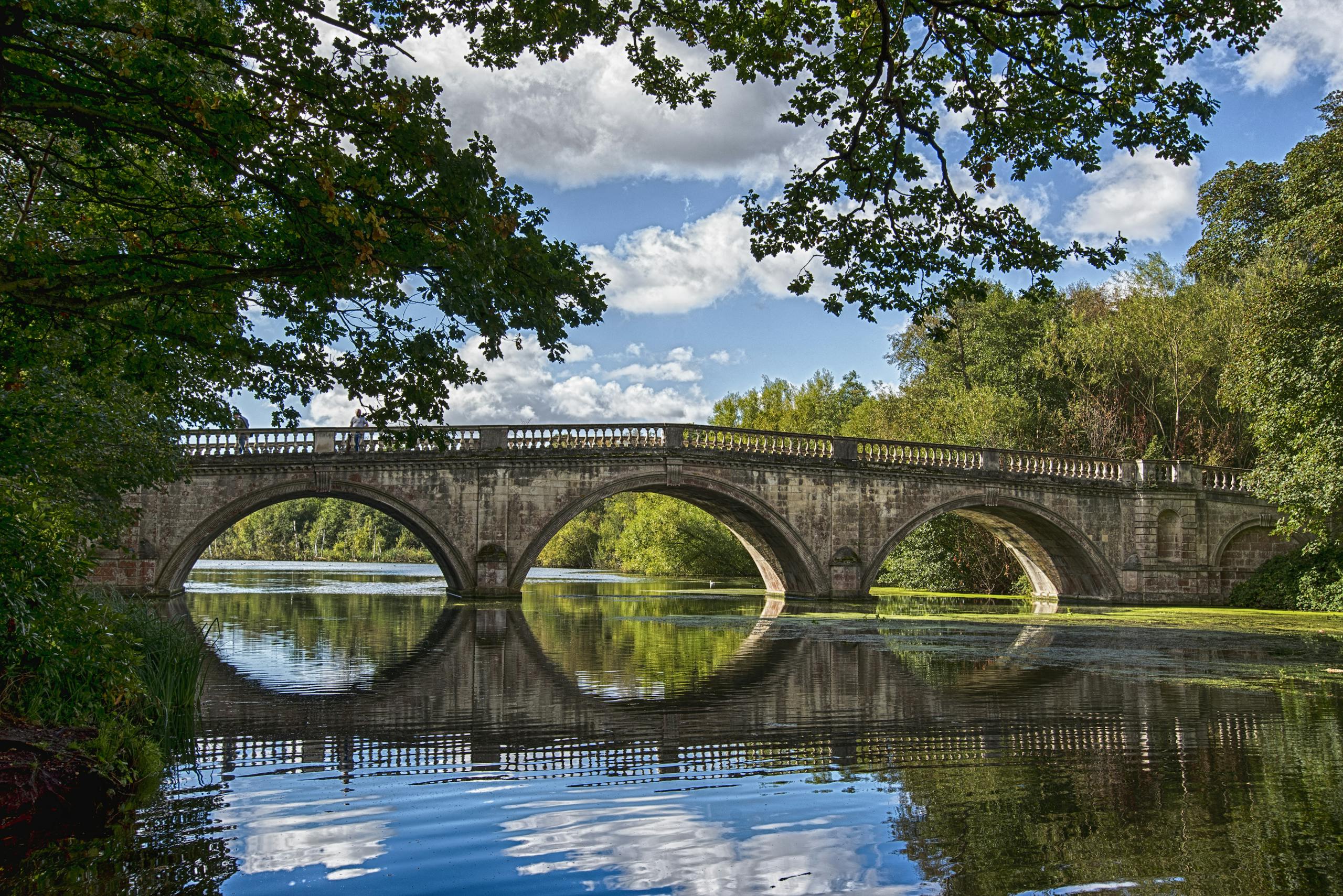 Ancient stone bridge arches over a calm river, surrounded by lush greenery and clear skies.